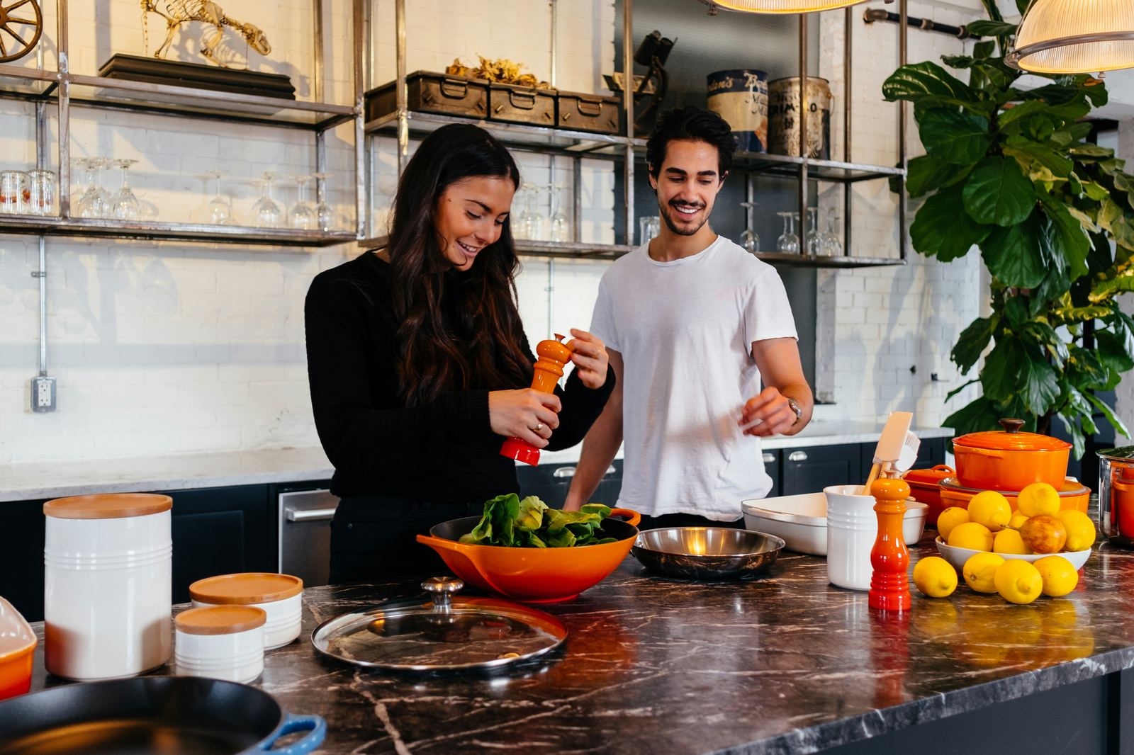 A couple in their kitchen prepping loose ingredients — lemons, greens, and pantry jars — on the counter before cooking